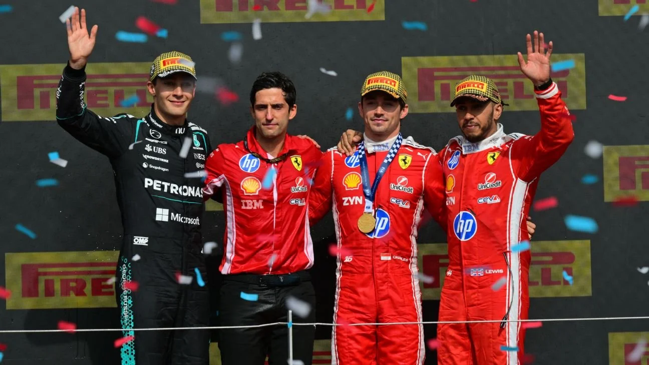 Four Formula 1 drivers stand on the winners' podium celebrating with confetti falling around them. The first-place driver is wearing a red Ferrari racing suit with a gold medal around his neck, and the third and fourth place drivers are also in red Ferrari suits. The second-place driver is in a black Mercedes racing suit. They are all waving and smiling.