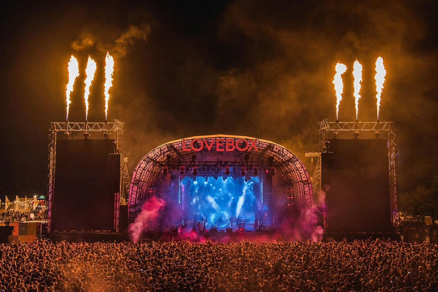 Nighttime outdoor music festival with a large crowd, stage with bright lights, fire effects, and the sign 'LOVEBOK' on top.
