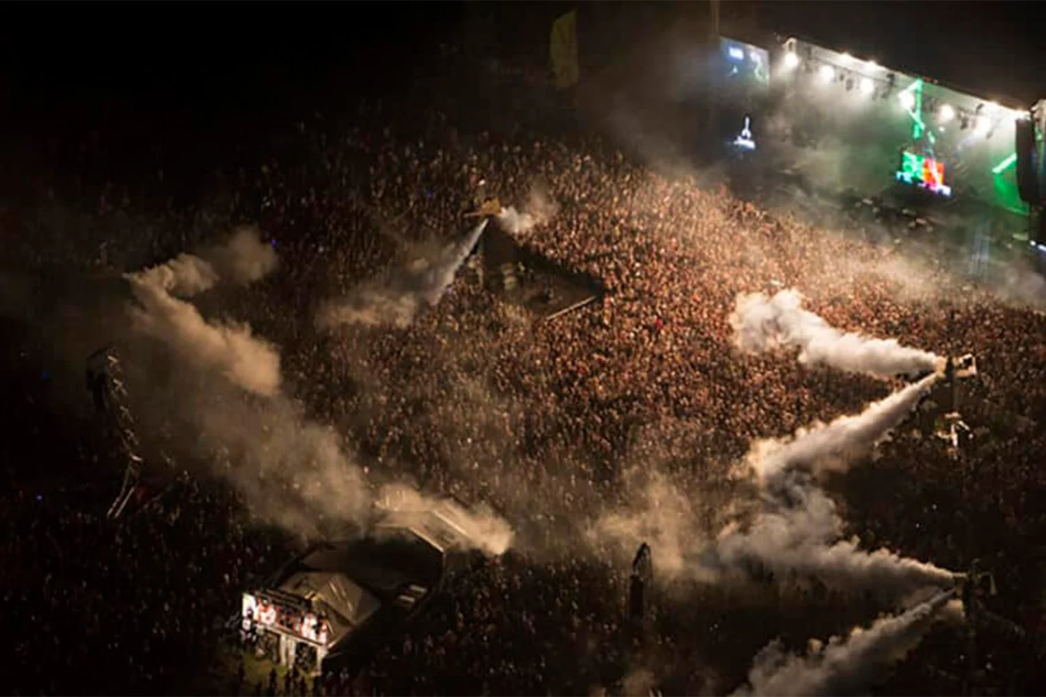 Aerial view of a large outdoor concert at night with a dense crowd, stage lights, and smoke effects.