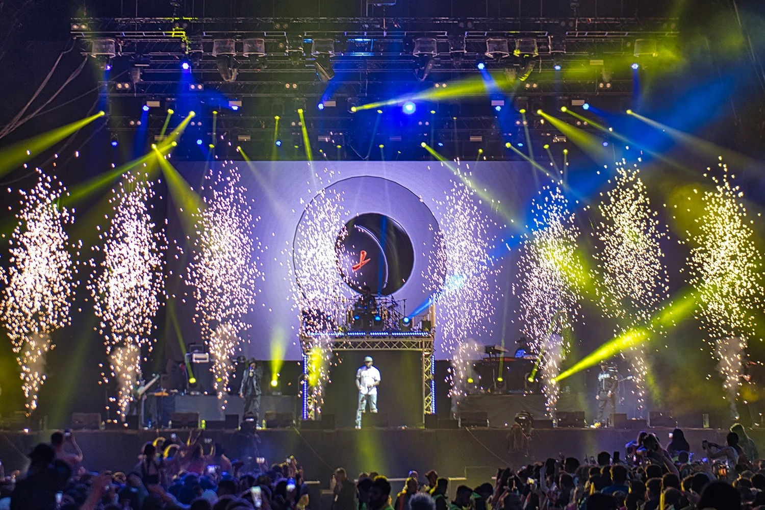 A live concert with a band performing on stage, surrounded by vibrant yellow and blue stage lighting, pyrotechnics, and fireworks, with an audience taking photos and watching.