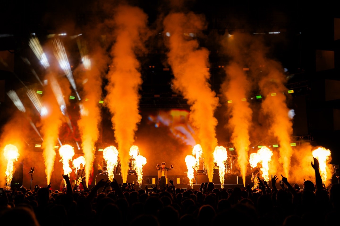 Live concert with pyrotechnics, fire, and smoke effects on stage, musicians performing, and audience with raised hands.