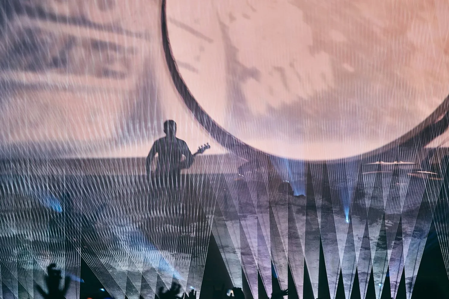 A musician playing guitar on stage with laser light displays and projections of a large moon in the background