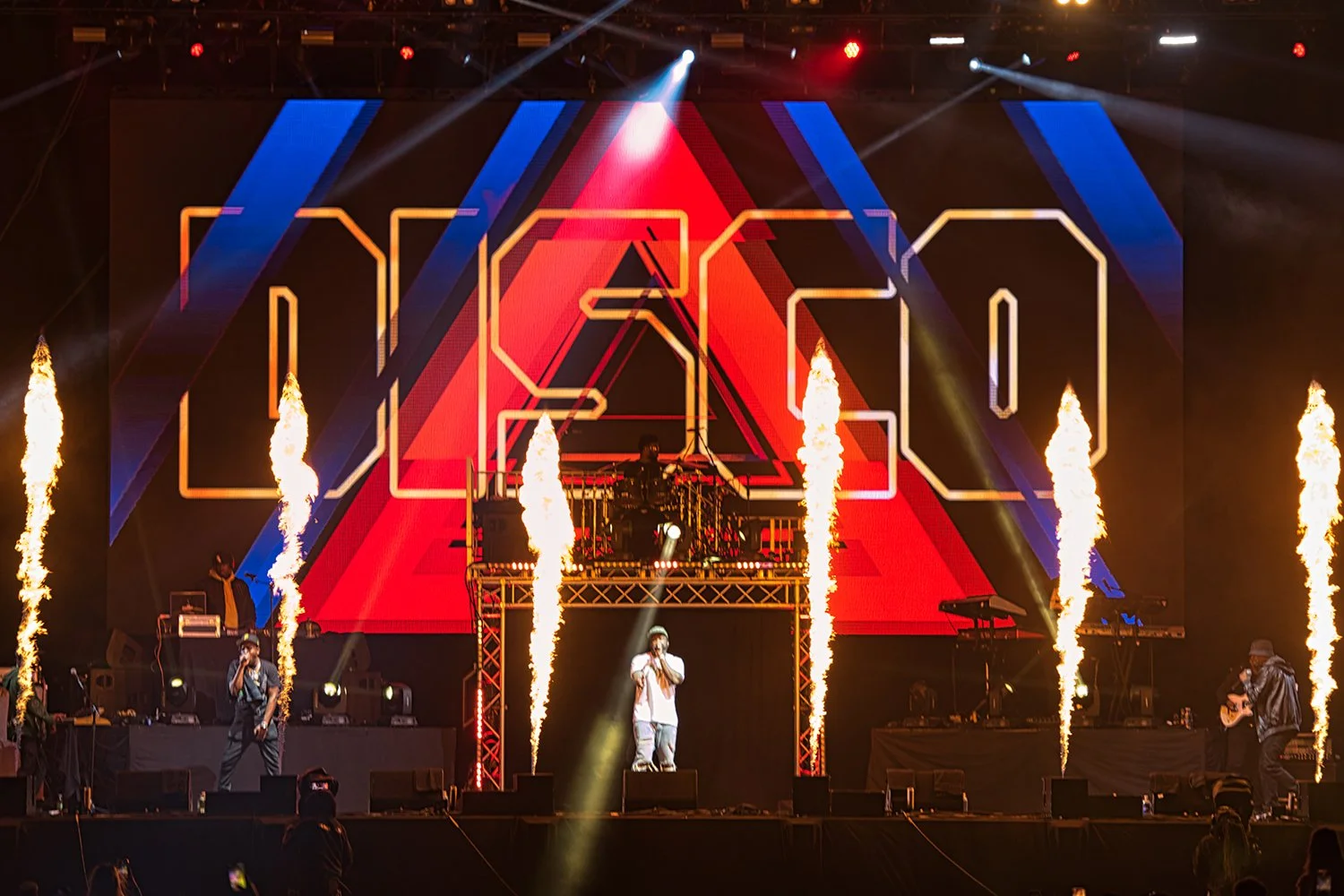 A live concert with band members performing on stage, large digital screen displaying