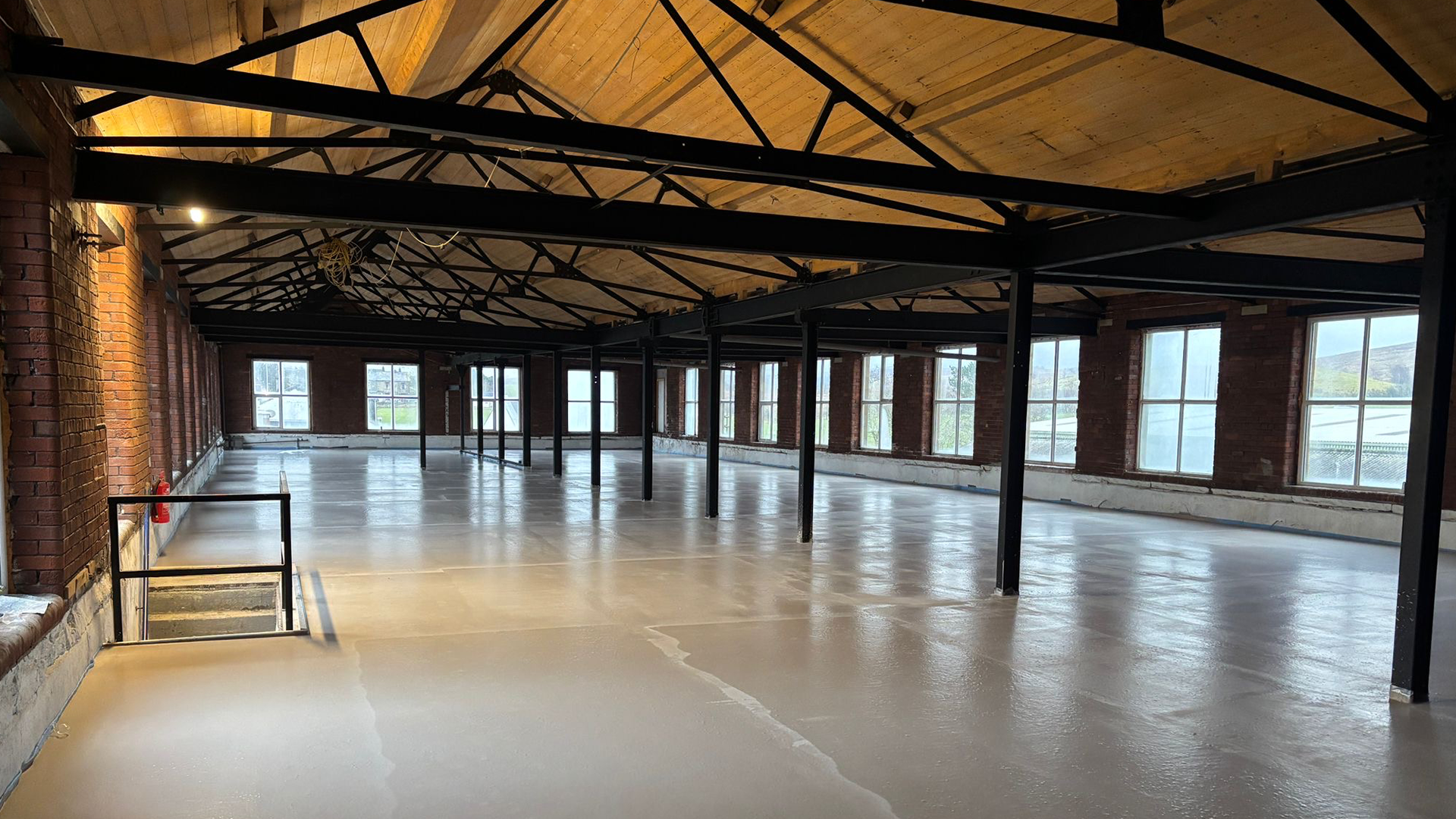Interior of a building under construction with brick walls, large windows, and a wooden ceiling supported by black metal beams.