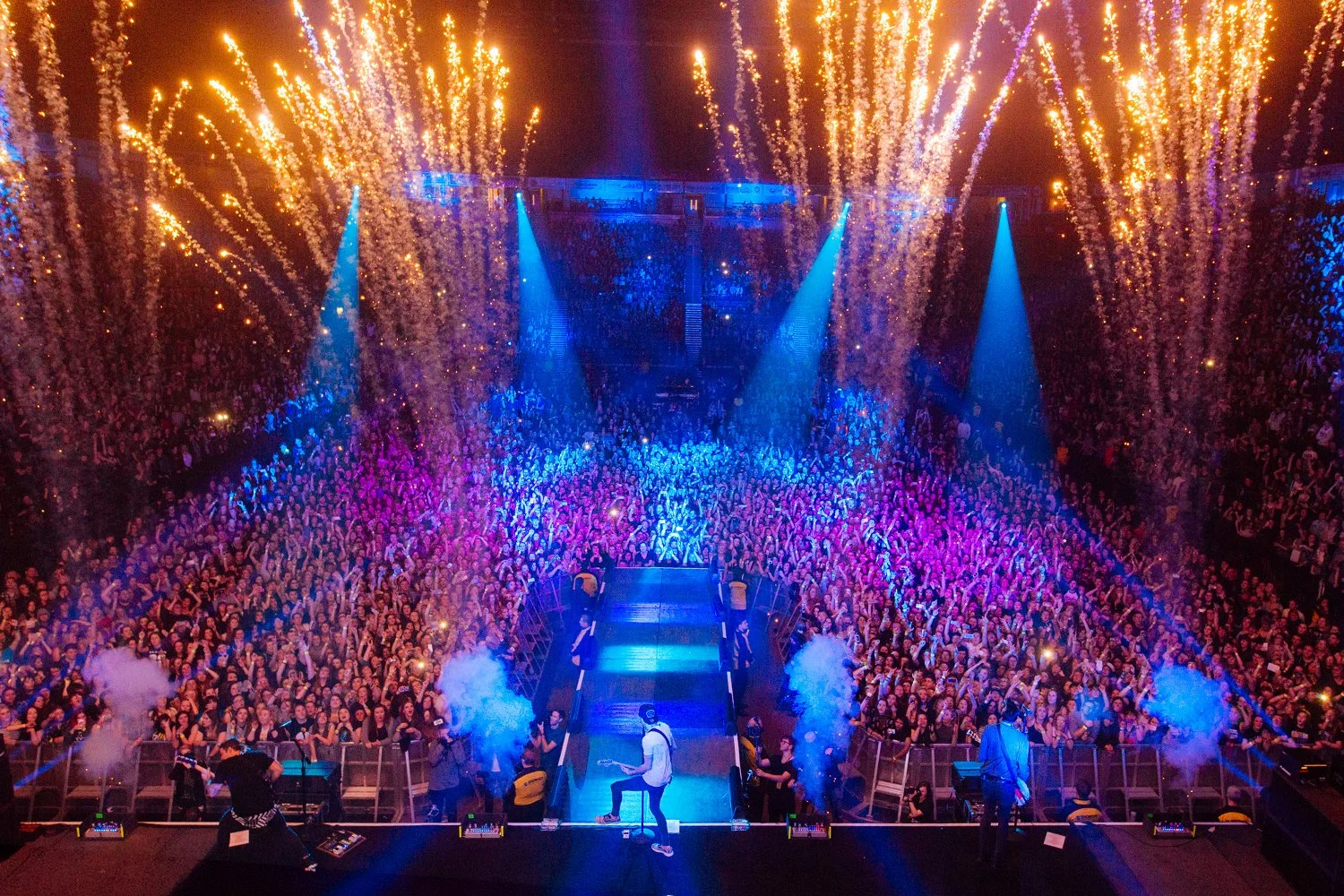 A concert hall filled with a large crowd of people, with purple, blue, and orange stage lighting, featuring a stage with musicians and special effects like smoke and fireworks.