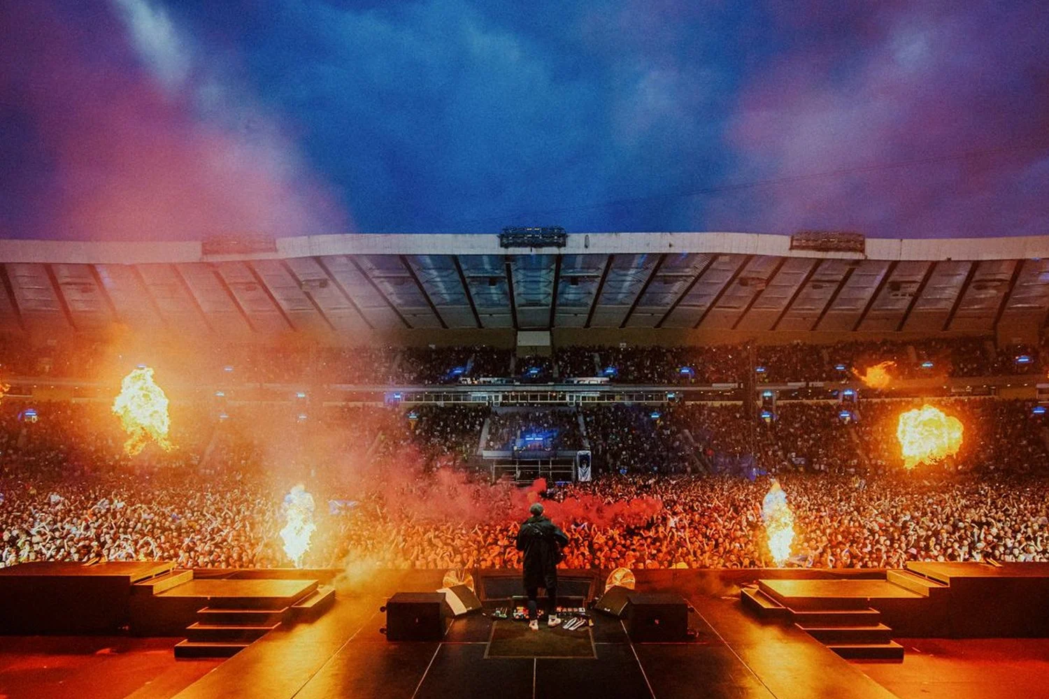 A DJ performs on stage at a large concert with flames and fire effects, a huge crowd in the stadium, and a dramatic cloudy sky overhead.
