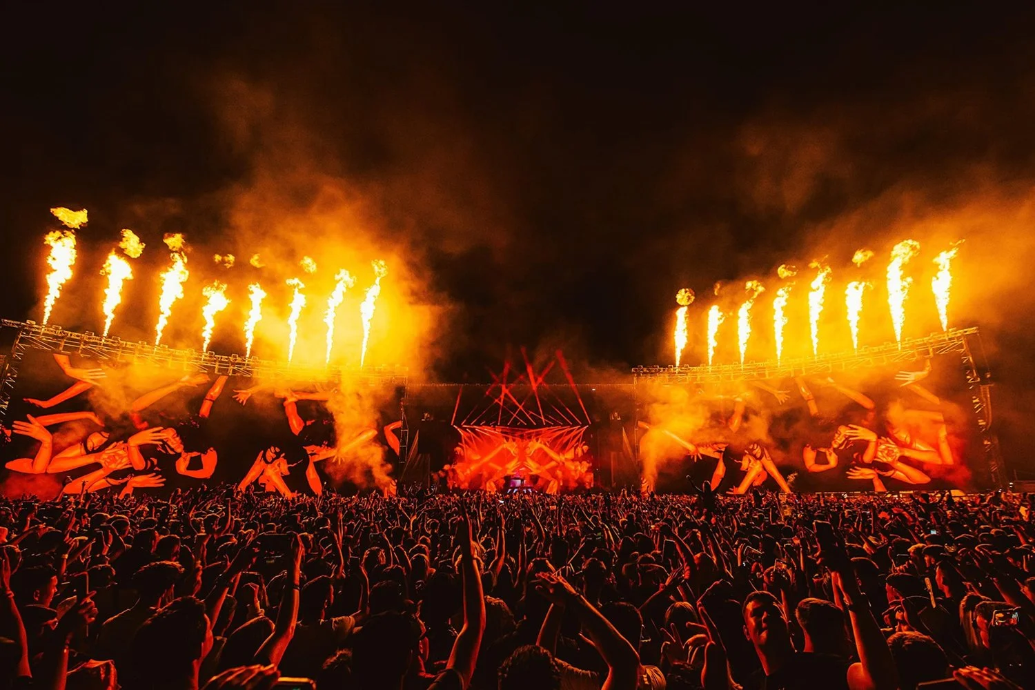 A large concert or music festival with a crowd of people, a stage with bright red lights, and pyrotechnics emitting flames against a dark night sky.