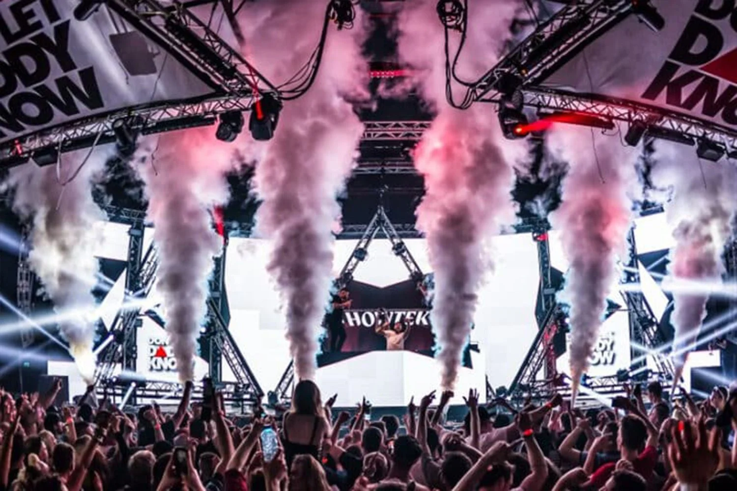 A large stage at a concert with smoke effects and multiple LED screens, with a crowd of people raising their hands and taking photos.