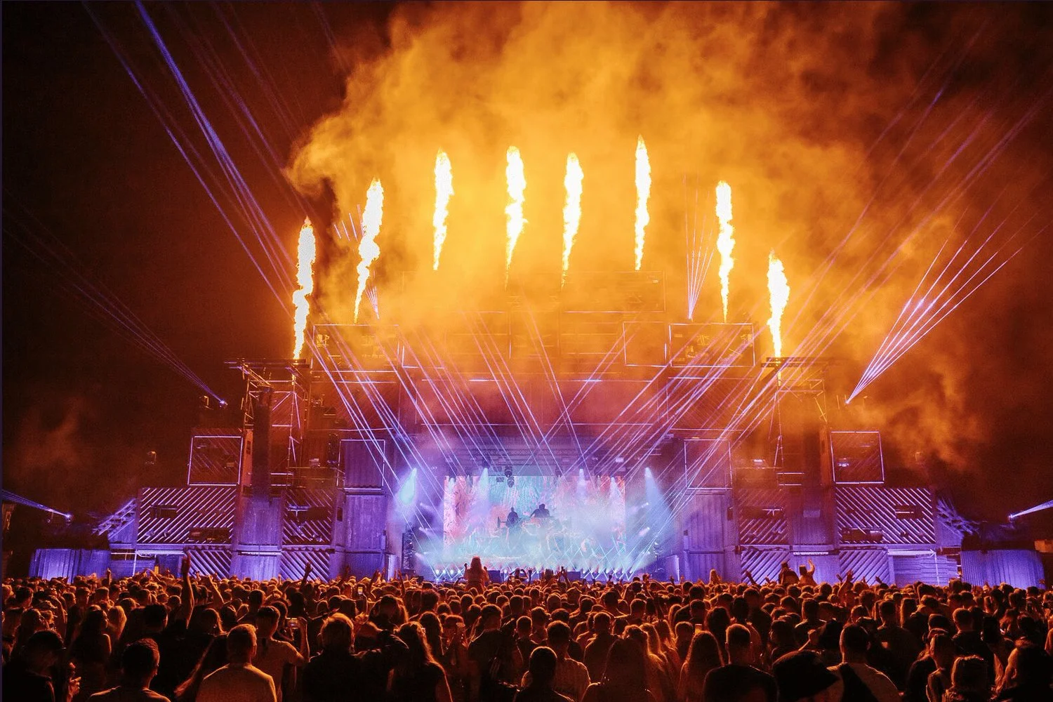 A large crowd watching a concert or festival at night with a stage featuring pyrotechnics, fire cannons, and colorful lighting effects.