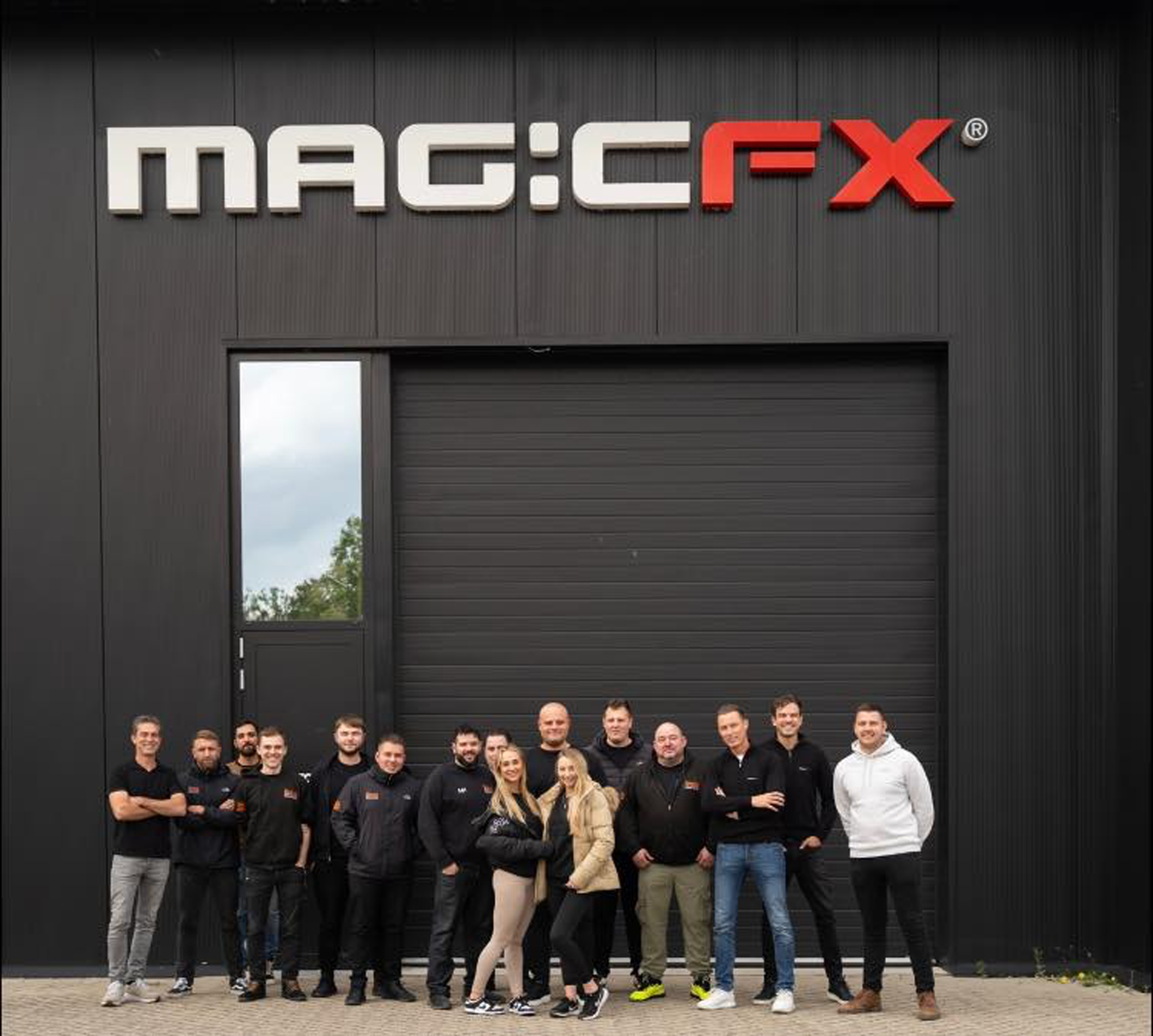 Group of people standing outdoors in front of a large dark building with the sign 'MACO.FX' on it.