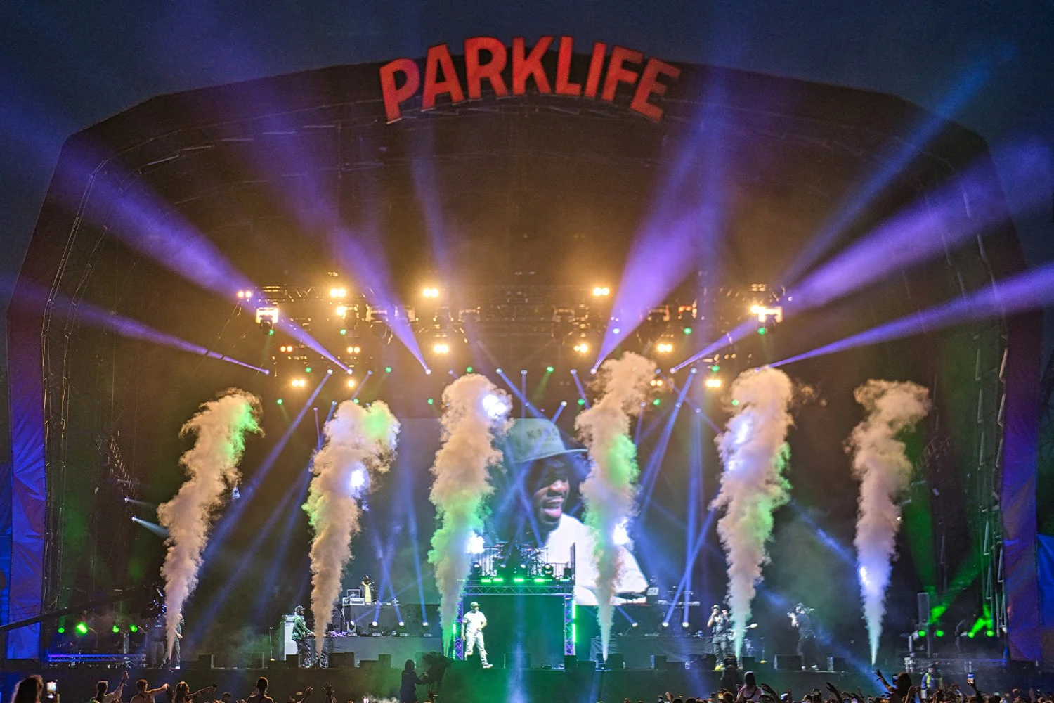 Concert stage with bright lights, smoke effects, and a large screen showing a performer wearing a white hoodie. The stage has a sign reading 'PARKLIFE' at the top, and audience members are visible in the foreground.