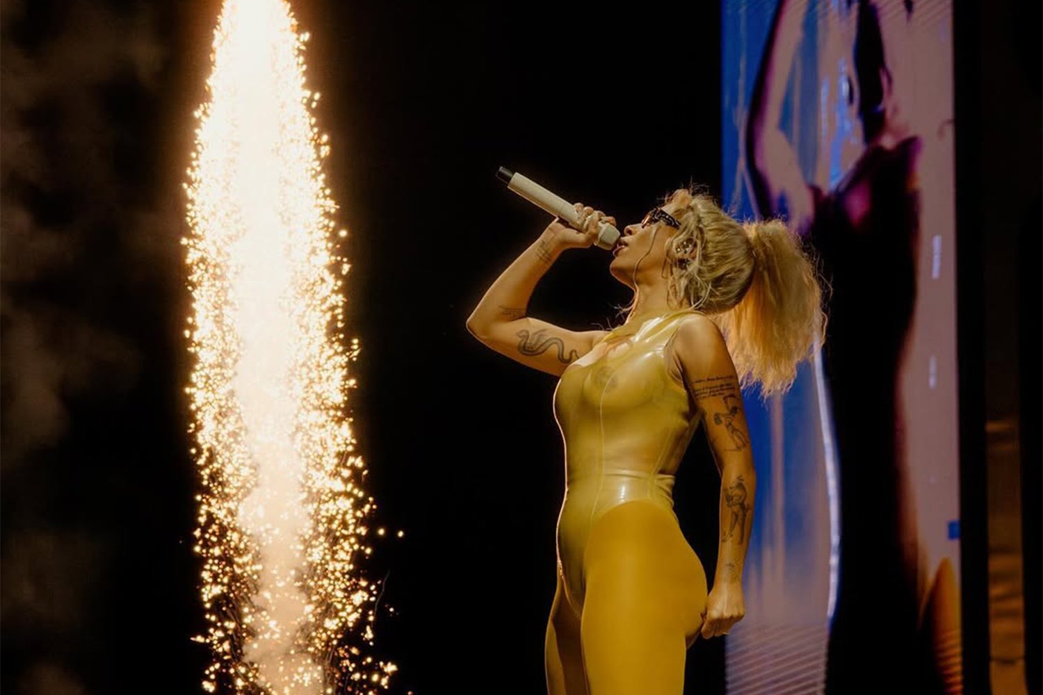 A woman known as a performer or singer, wearing a shiny yellow bodysuit, singing into a microphone on stage with fireworks in the background.