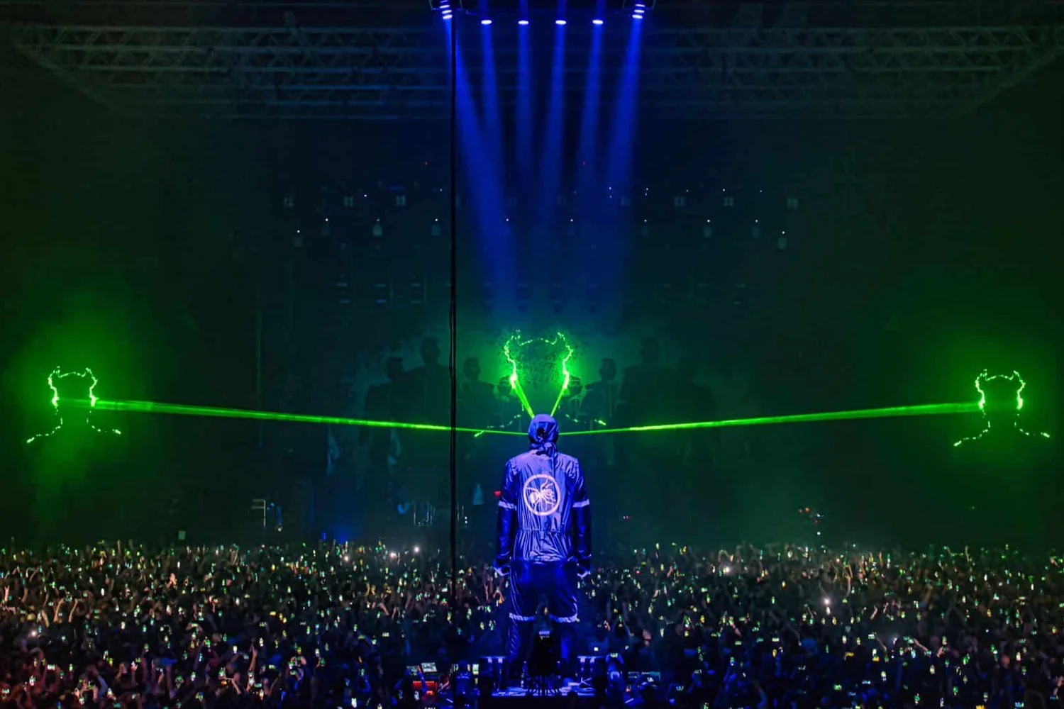 A concert scene featuring a performer on stage dressed in a dark jacket with a logo on the back, with green laser lights and a large crowd holding up lights or phones. The background includes blue stage lights and silhouettes of other performers or audience members.