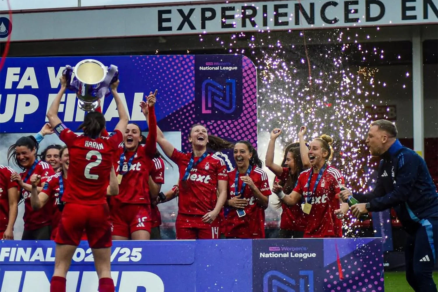 Women soccer players celebrating with a trophy on a stage, surrounded by confetti and cheering with medals around their necks.