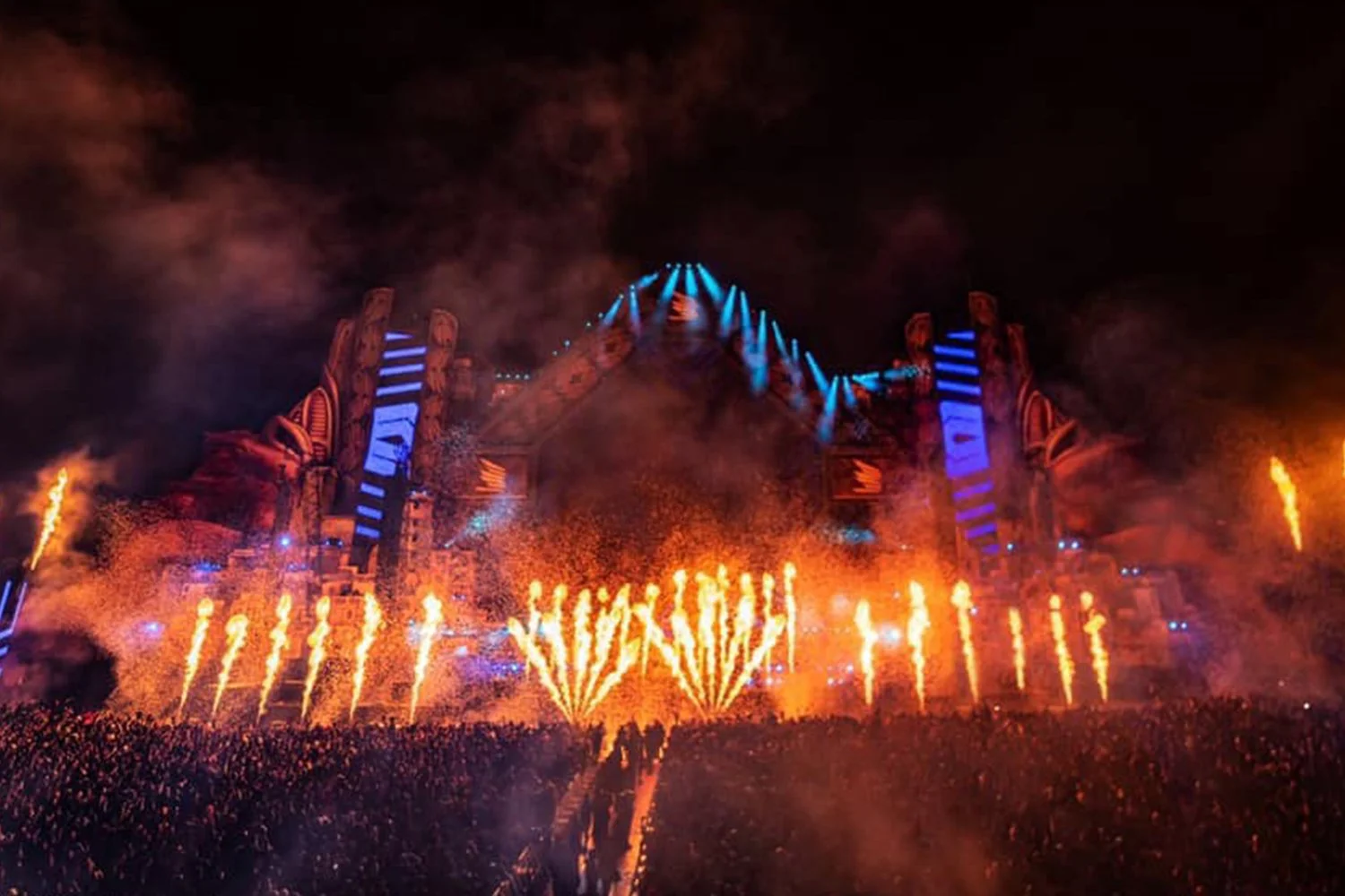 Nighttime music festival stage with blue and orange lights, pyrotechnics, and a large crowd.