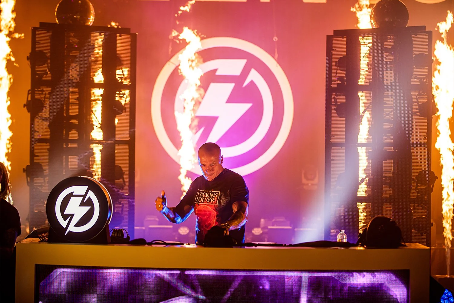 A DJ performer's set with fire effects, behind a large circular logo with a lightning bolt inside a circle, at a music event. The background features two large LED screens and flames on either side.