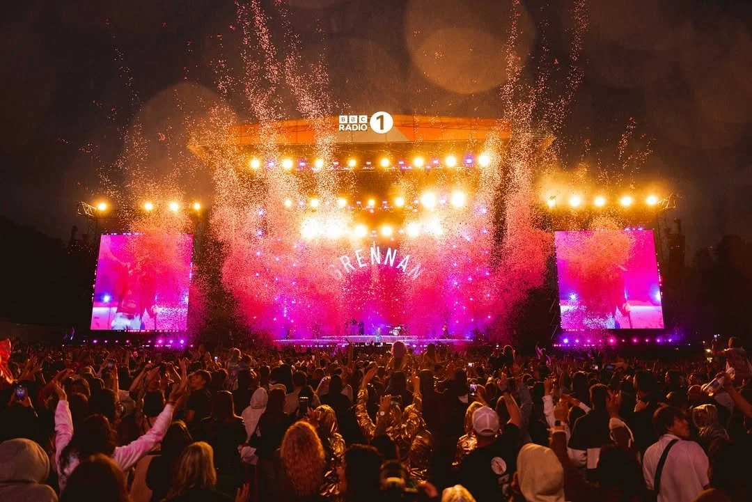 Crowd at a music festival with a stage lit up by colorful lights and pyrotechnics, and large screens displaying performers. The name 'Drennan' is visible on the stage background.