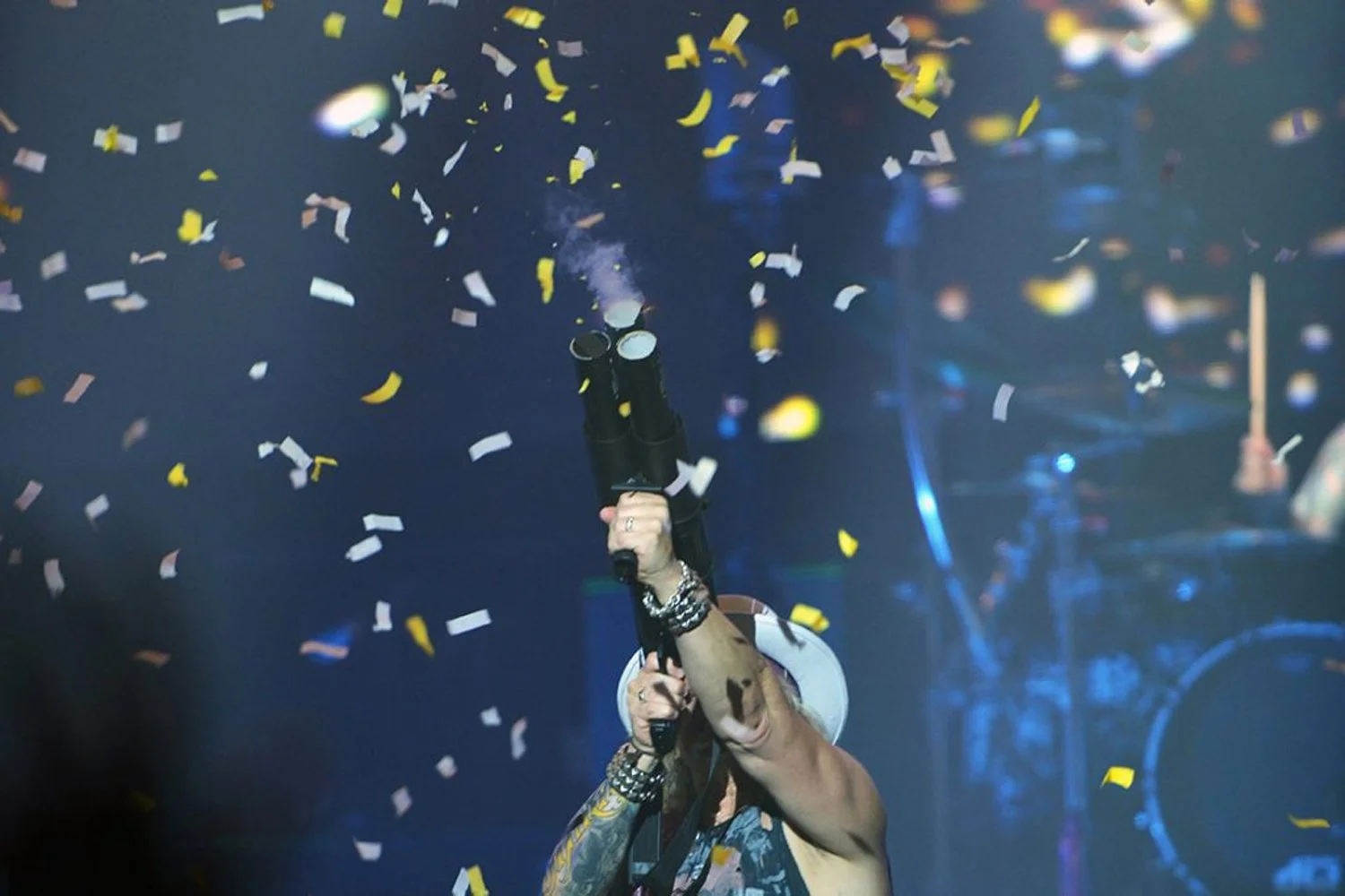 A person wearing a hat and multiple bracelets holds a confetti cannon pointed upward as yellow and white confetti falls around them during a celebration or concert.