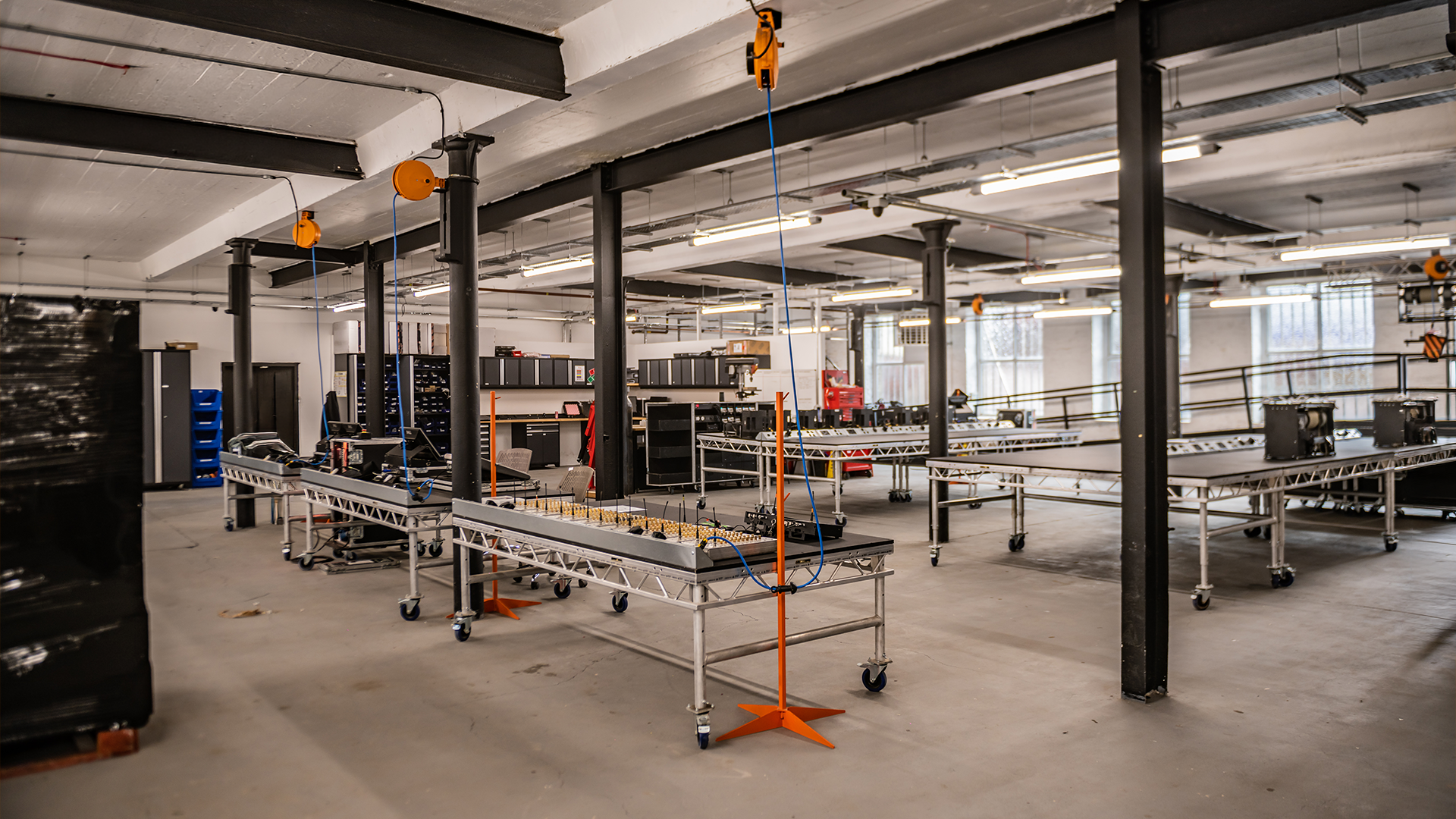 Industrial workspace with metal workbenches, tools, and orange lifting straps, illuminated by fluorescent lights, with large windows and structural beams.
