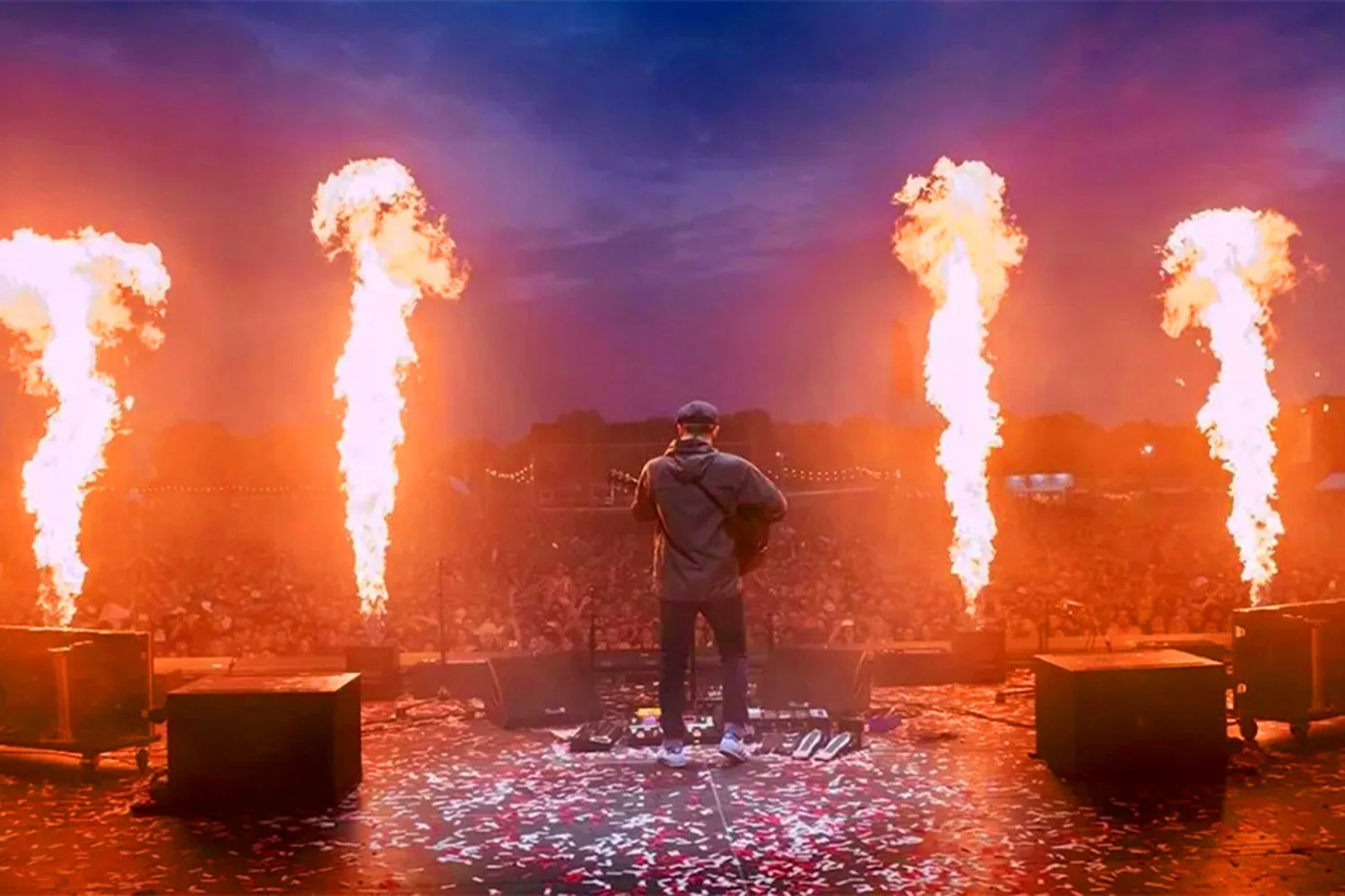 A musician performing on stage with fire pyrotechnics shooting up along the sides, a large crowd in front, and confetti on the ground.