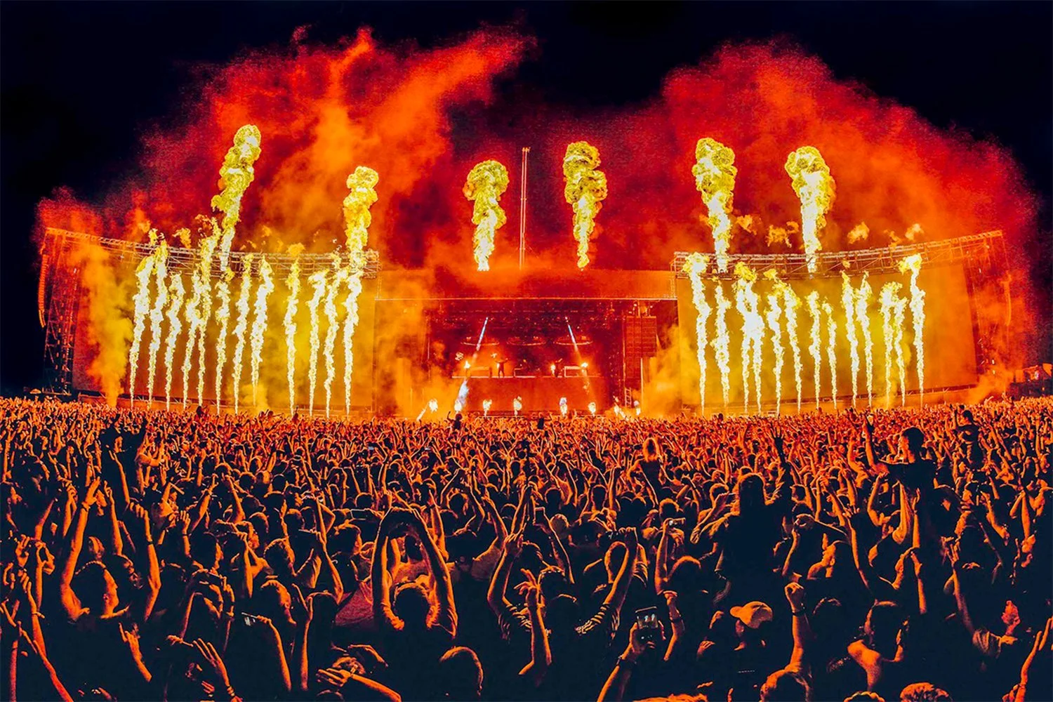 A large outdoor concert at night with a crowd of people raising their hands and cheering. The stage has pyrotechnics with multiple flames shooting upward, creating a fiery display.
