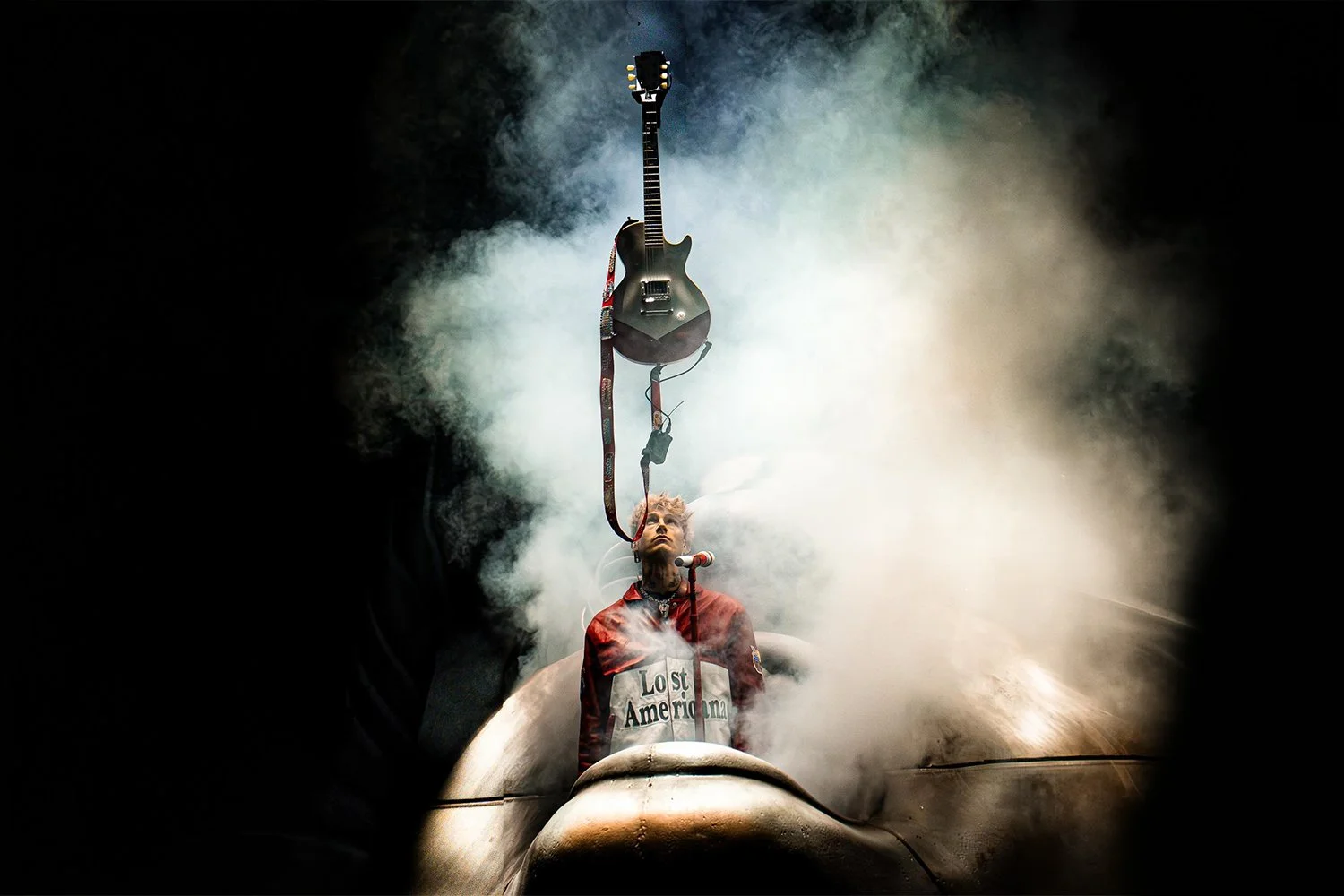 A person with light-colored hair and tattoos on their neck, standing behind a microphone, wearing a red jacket with white and black text, with a smoke-filled background. There is an electric guitar hanging directly above their head, suspended in midair.