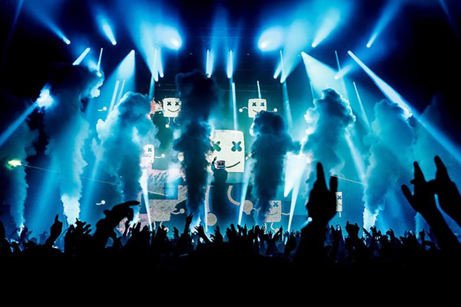 Concert scene with bright blue and white stage lights, smoke effects, moving hands in the audience, and digital screens displaying cartoonish smiling robot faces with crosses for eyes.