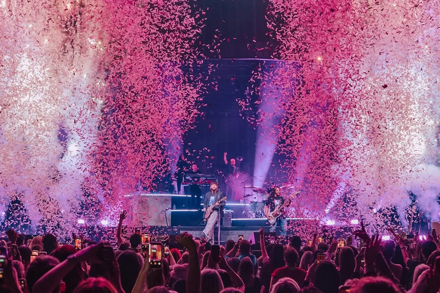 A large crowd at a concert with multiple performers on stage, surrounded by pink and purple confetti, sequins, and stage lighting.