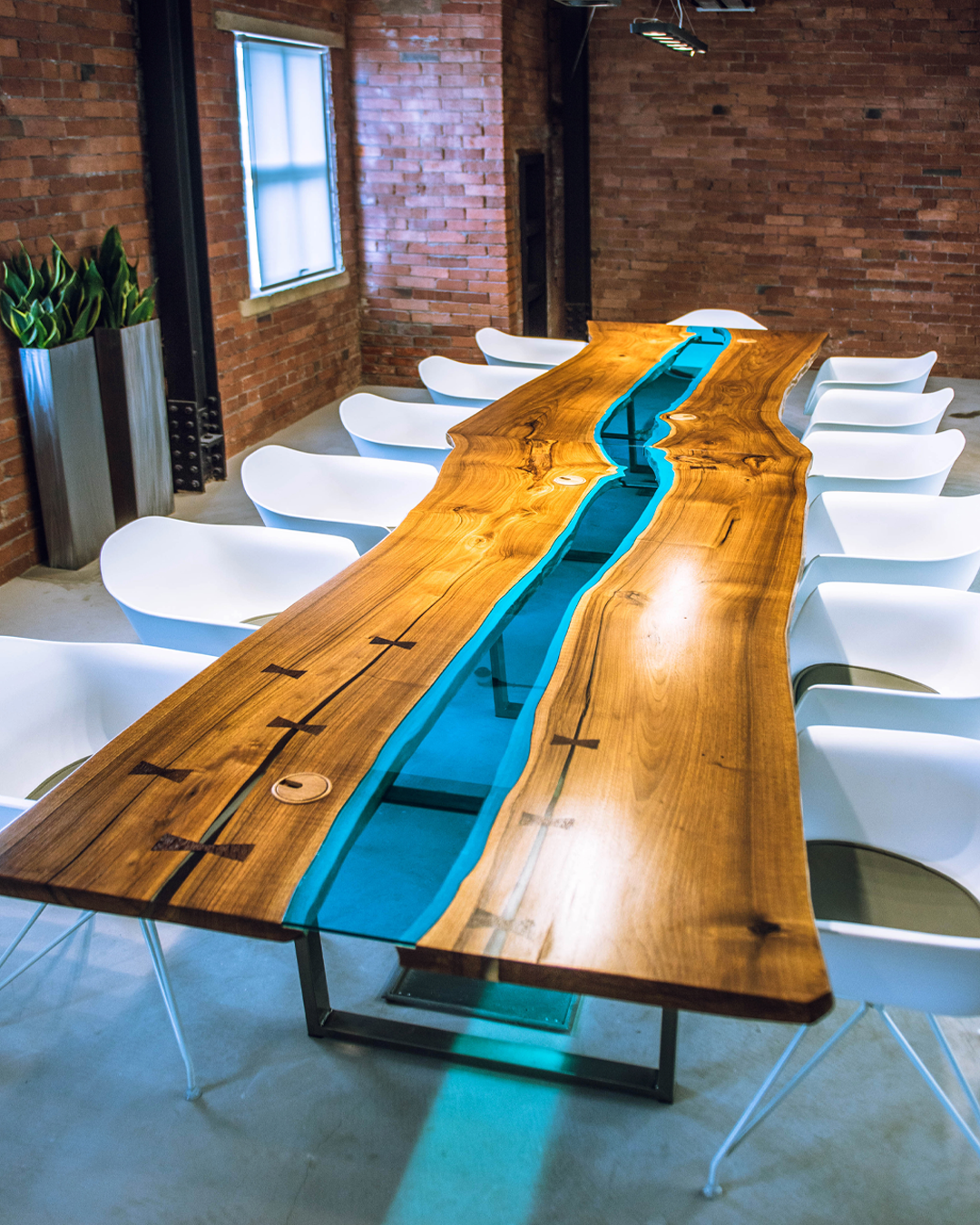 A large wooden conference table with a blue resin river in the middle, surrounded by white modern chairs, in a room with exposed brick walls and a window.