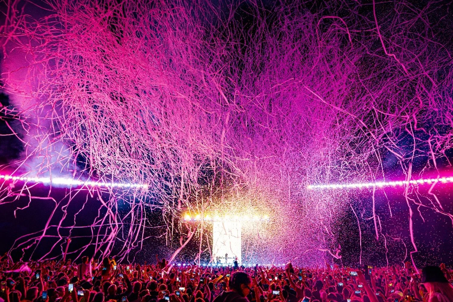 Large crowd at a concert or festival with pink and purple streamers and lights in the night sky.