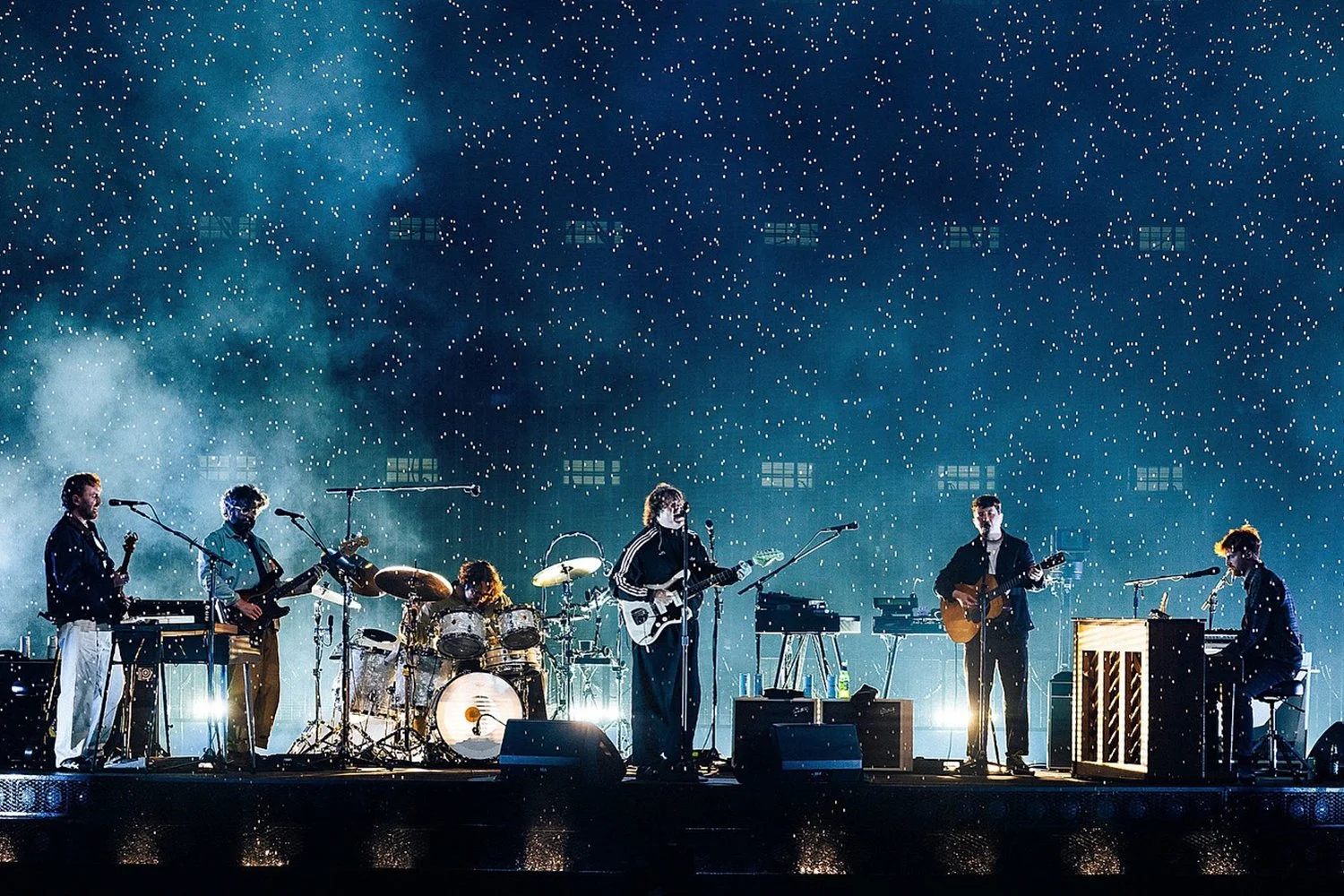 A band performing on stage with a starry background and smoke effects, featuring a keyboardist, guitarist, drummer, and vocalist.