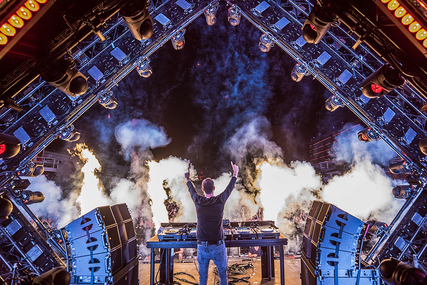 DJ performing on stage at a music concert, with smoke effects and stage lighting, under a canopy of stage lights.