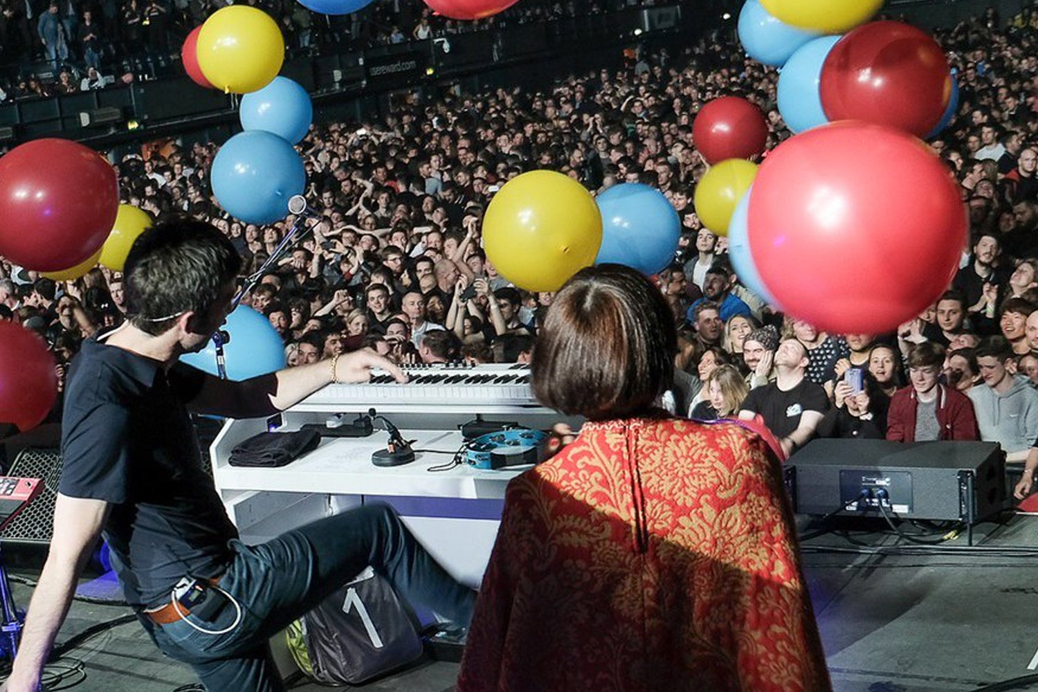 Photo of musicians performing on stage at a concert with a large audience. The stage has colorful balloons hanging above, and the crowd is excited and engaged.