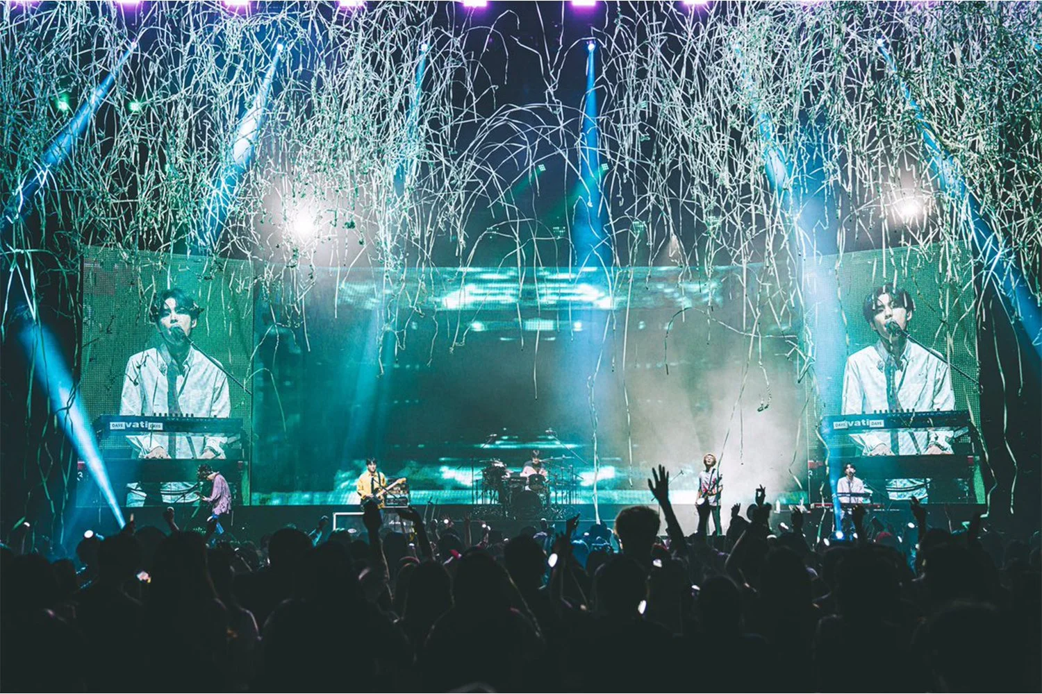 A live concert features a band performing on stage with large screens. The stage is decorated with hanging streamers and colorful lights, with an audience in the foreground cheering and raising their hands.