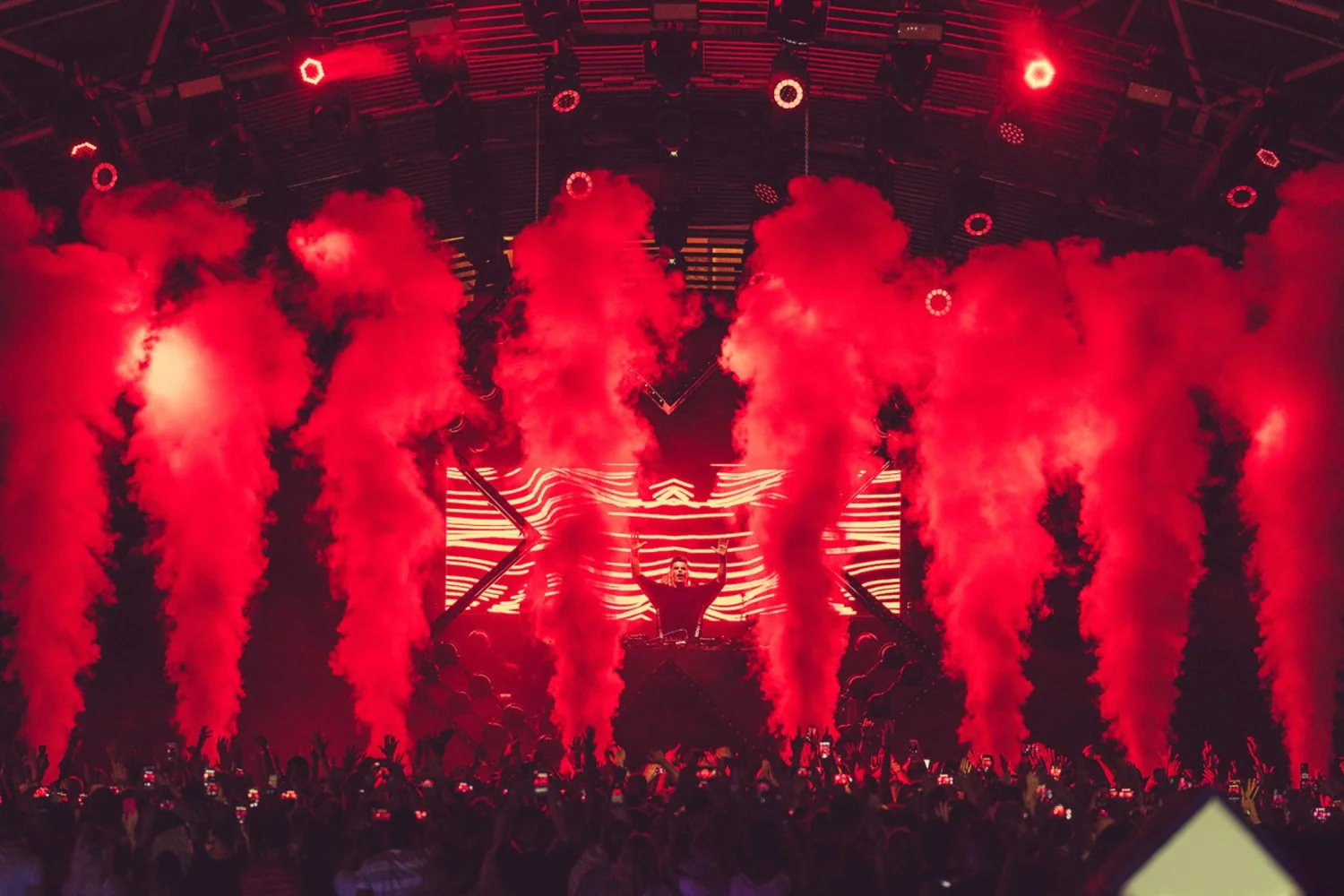 A DJ performs on stage surrounded by red smoke and LED screens, with a large crowd in front capturing the show with their phones.