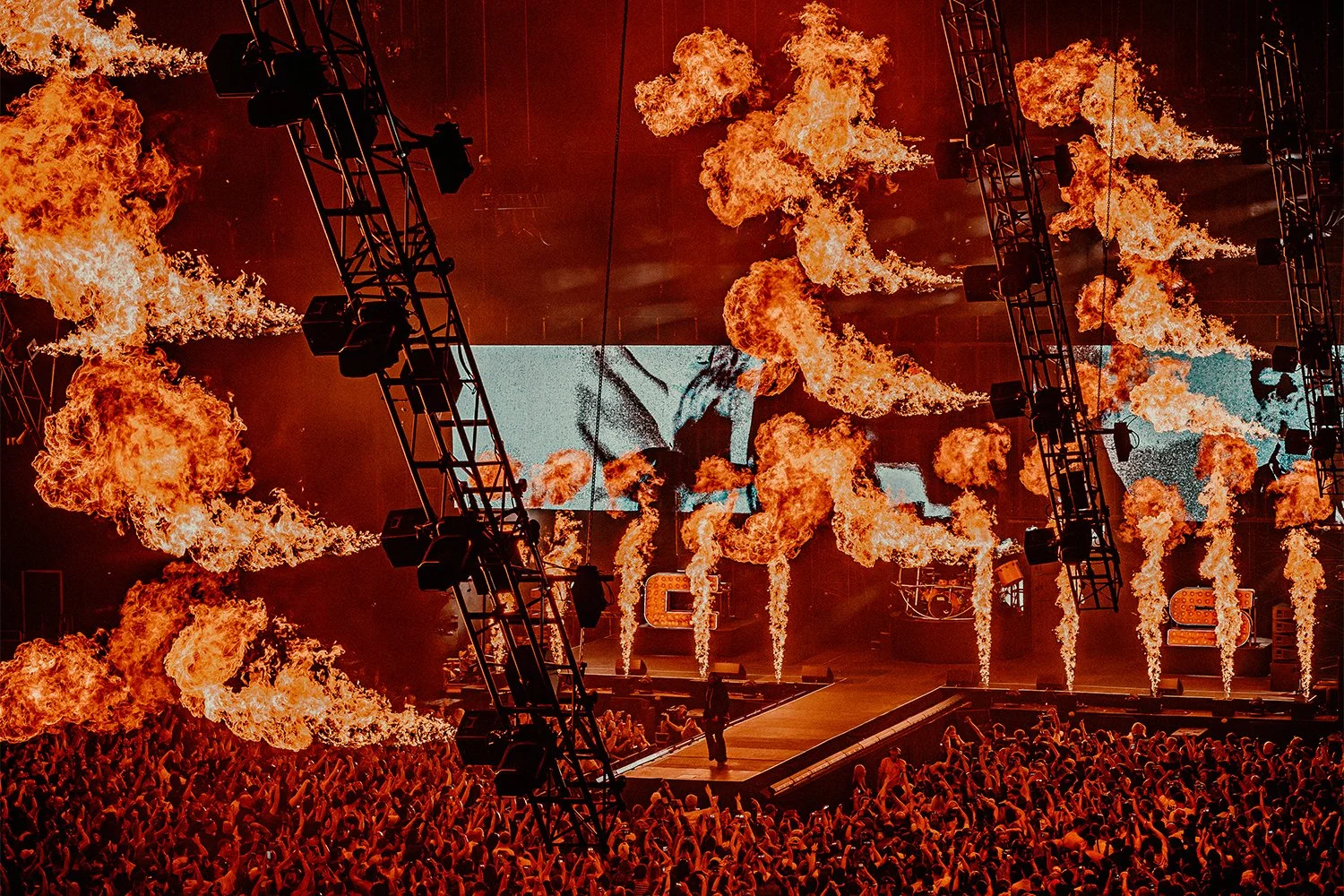 A concert stage with large flames shooting up from the sides, a DJ or performer silhouette on a platform, surrounded by a large crowd of fans.