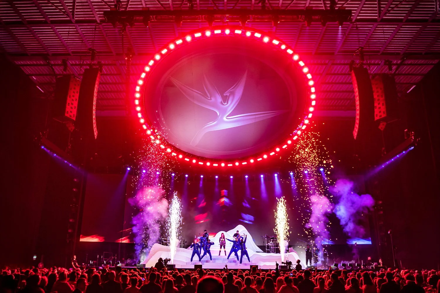 A large indoor concert with a stage, a band performing, colorful lighting, fireworks, and an audience in the foreground. The ceiling has a circular LED display with a logo.