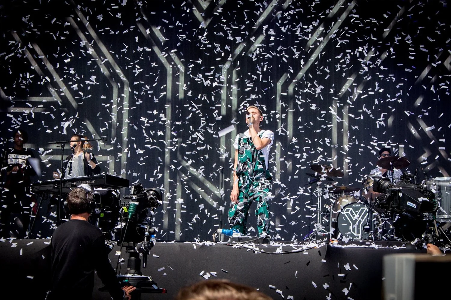 Live concert with a band performing on stage, confetti falling, and audience members capturing the moment.