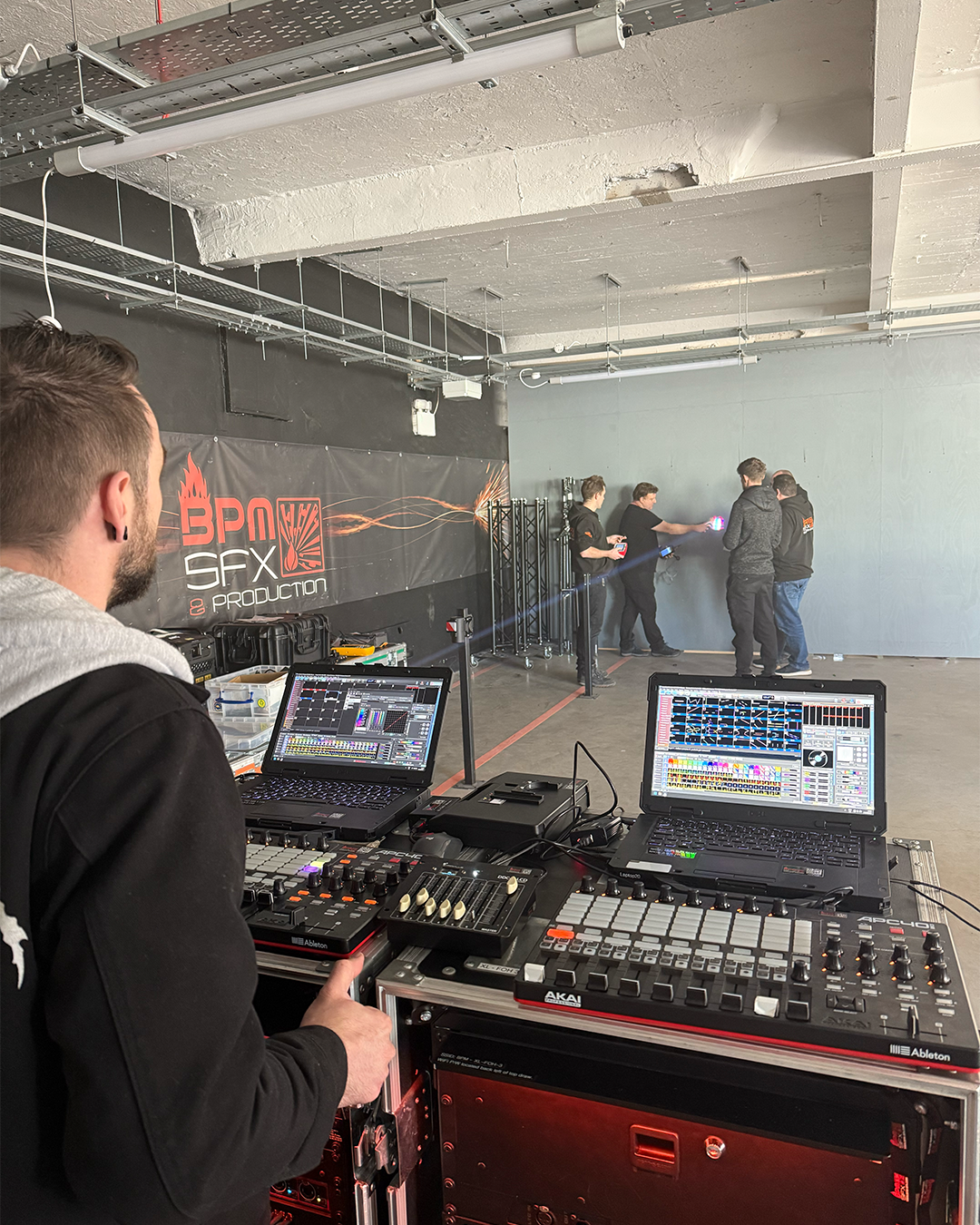 A sound engineer operates DJ equipment in a warehouse, preparing for a stage setup. In the background, four people are testing lighting and visual effects on a plain wall.