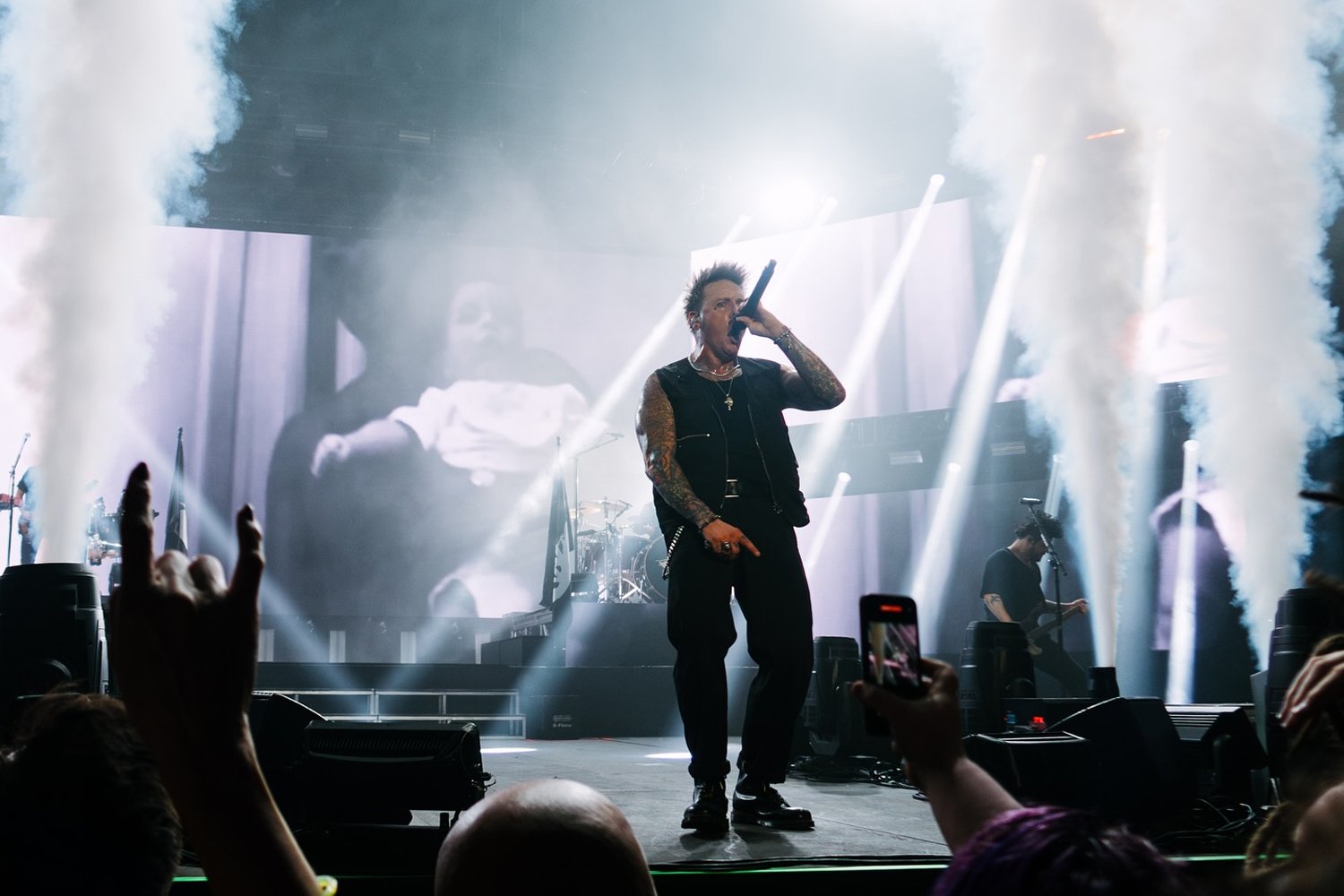 A male performer with tattoos sings into a microphone on stage during a concert, with a drum set and band members in the background, surrounded by stage smoke and bright lighting.