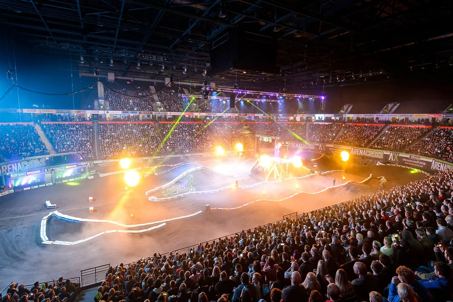 Indoor arena with a dirt motocross track, bright stadium lights, colorful laser lights, and a large audience watching an off-road motorcycle race.