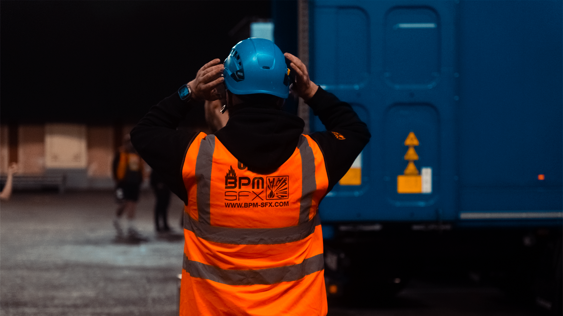 A worker wearing a blue safety helmet and an orange reflective vest stands in front of a large blue truck, with hands adjusting the helmet. The back of the vest features a logo and a website.
