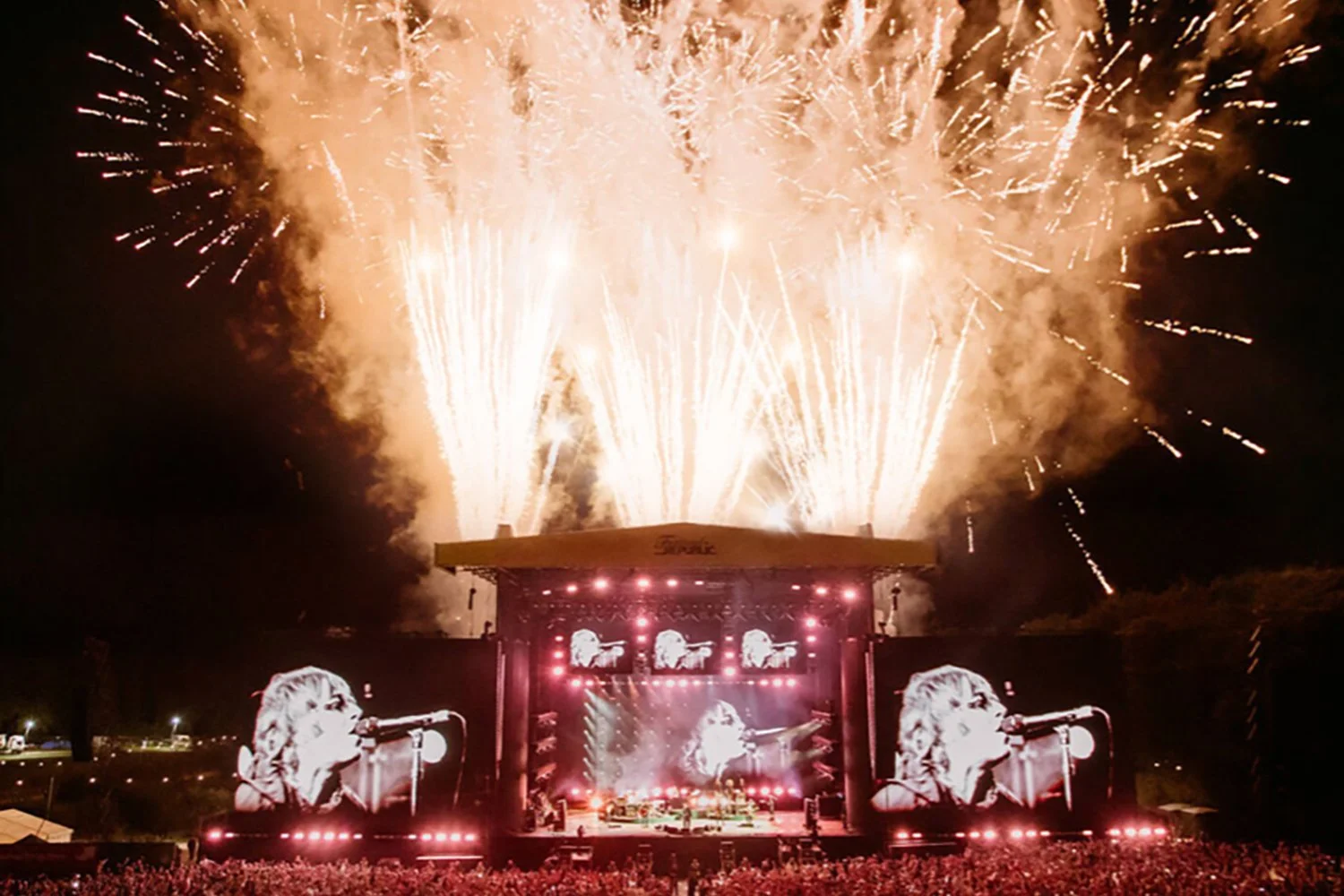 A concert stage with large screens showing a female singer, and fireworks exploding in the night sky above.