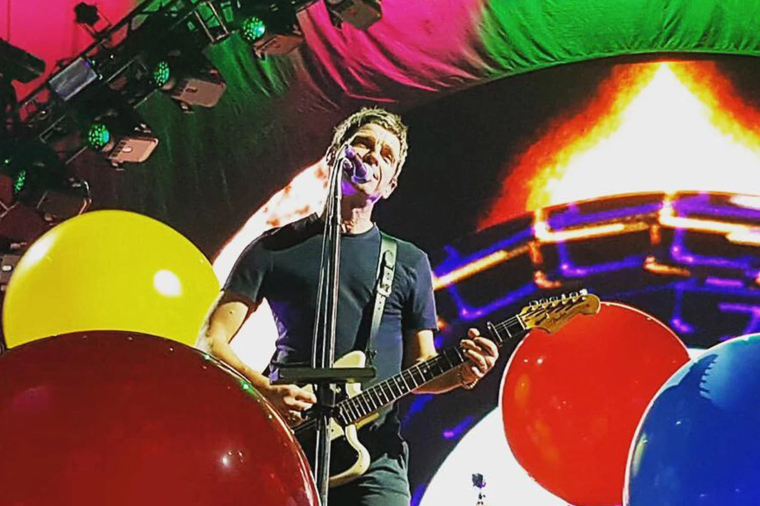 A man playing an electric guitar on stage surrounded by colorful balloons and vibrant background lights.
