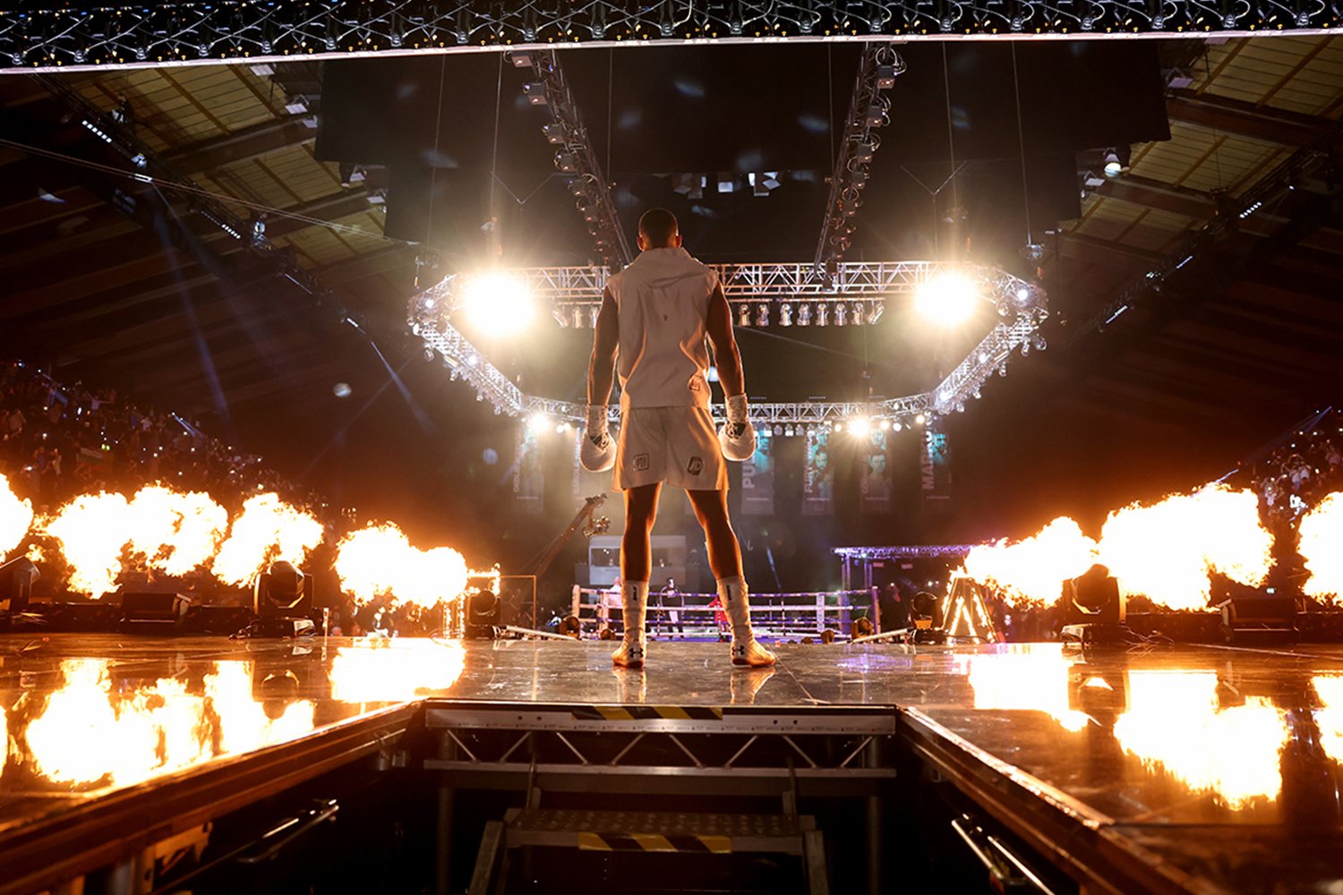 A boxer standing in a boxing ring with flames surrounding him, bright lights above, and a cheering crowd in the background.