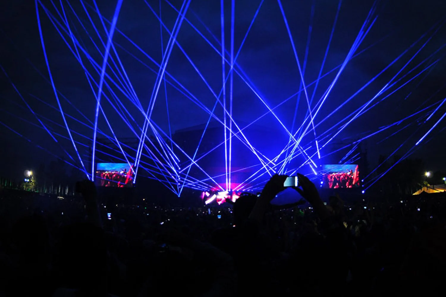 Nighttime concert with blue laser lights crisscrossing above a large crowd, two large screens displaying the event, and silhouetted audience members taking photos.