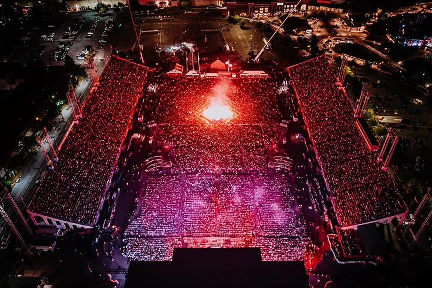 Aerial view of an outdoor concert or event at night with a large crowd gathering around a lit stage, illuminated in red and purple lighting.
