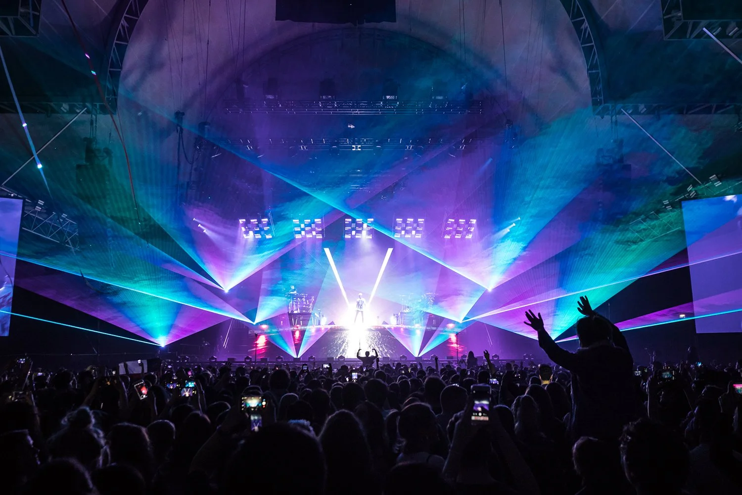 A concert with a light show featuring colorful lasers and bright stage lights, audience members capturing the event on their phones, and a performer on stage.