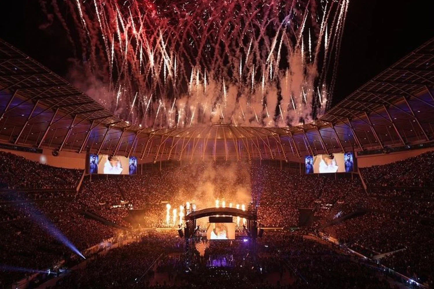 Fireworks exploding above a stadium during a large event, with spectators and large screens displaying images.