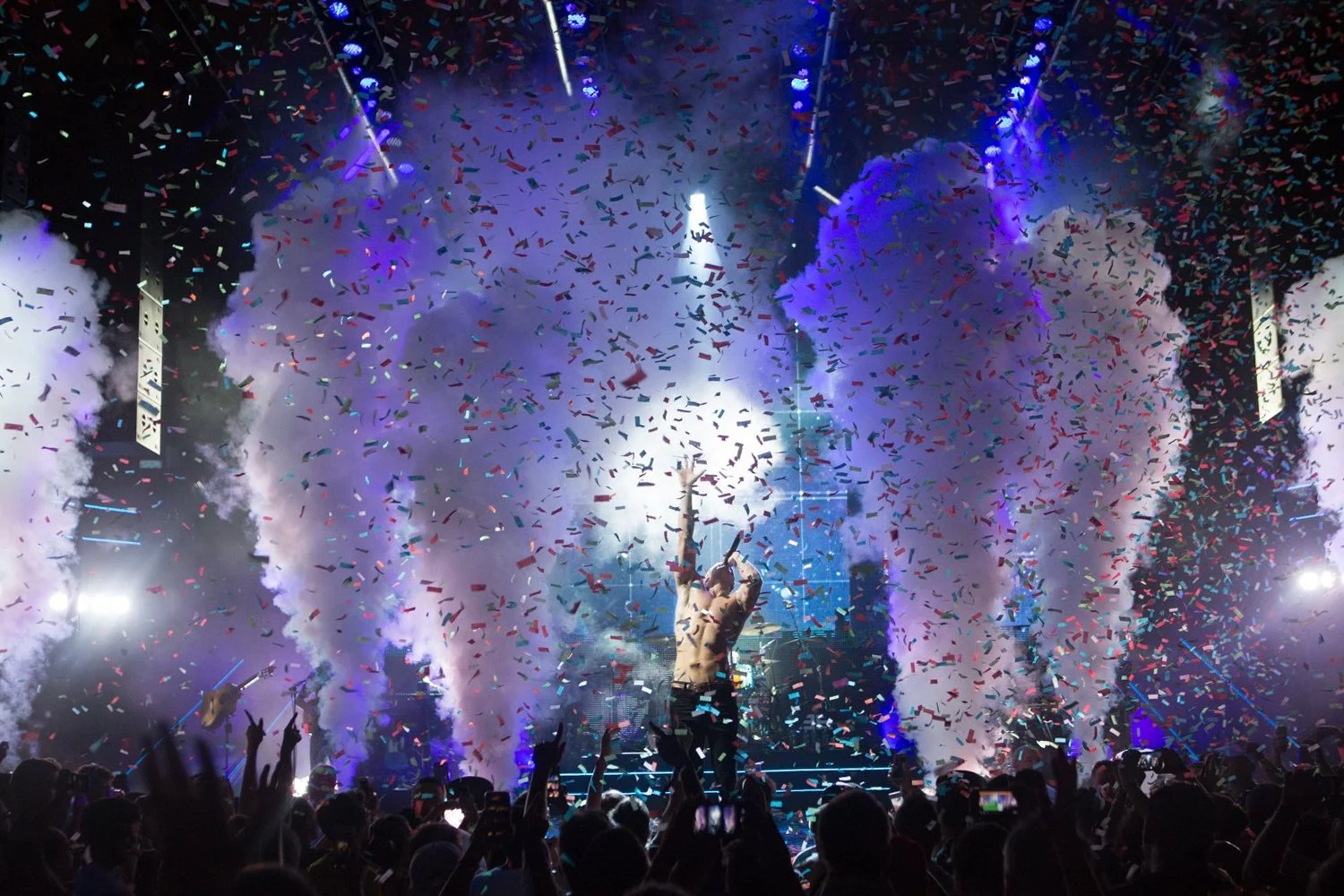 A concert scene with a performer on stage surrounded by colorful confetti and smoke effects, with an audience watching and taking photos.